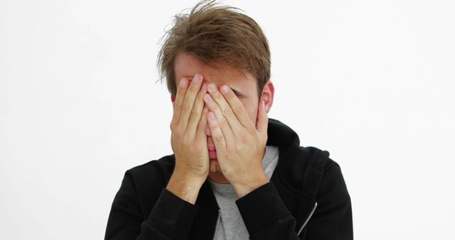 This image of a young man with his face covered by his hands conveys feelings of distress, anxiety, and frustration. It can be used for articles or campaigns focusing on mental health, stress management, or conveying emotional and psychological states. Ideal for illustrating the impact of overwhelming situations or personal struggles.
