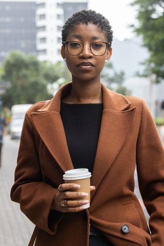 Confident black professional woman holding coffee on city street in brown coat and glasses