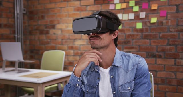 A young man in a denim shirt is exploring virtual reality using a VR headset in a modern, creative office featuring a brick wall and colorful sticky notes. Ideal for depicting innovative workplace environments, tech advancements, remote work trends, or creative industry settings.