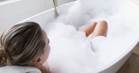 Woman relaxing in bubble bath in minimalist freestanding tub with brass faucet, soft light