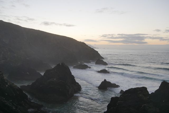 Misty cliffs overlook waves receding at dawn. The rocky coastal landscape and calm ocean make this scene perfect for depicting tranquility, nature's beauty, and peaceful moments. Ideal for use in travel brochures, nature magazines, and blogs focusing on coastal scenery.