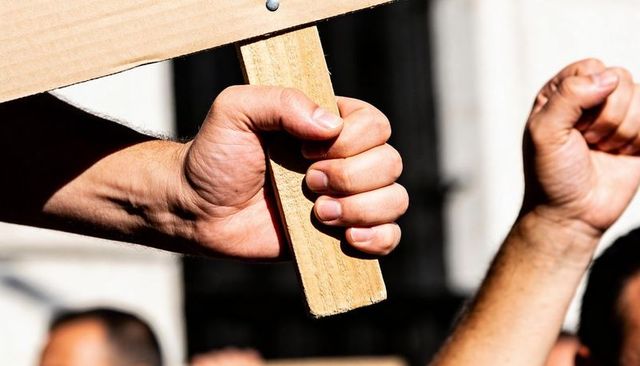 Sunlit hand gripping wooden protest sign handle with raised fists in crowd, closeup