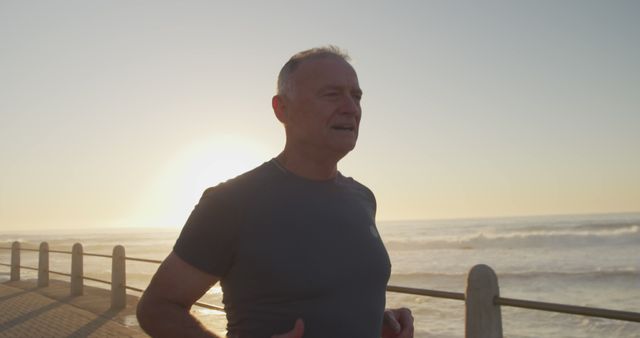 Senior man jogging along a seaside promenade during a beautiful sunset. This image is ideal for illustrating themes related to fitness, health, wellness, active lifestyles, and outdoor exercise. Perfect for use in health and fitness blogs, senior lifestyle articles, workout apps, and wellness advertisements.