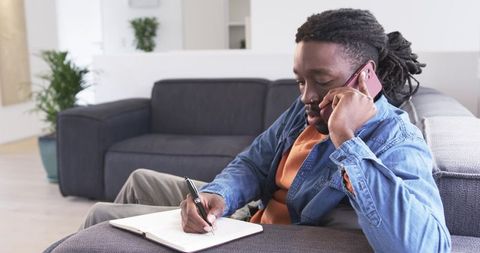 African american man taking notes during phone call on sofa, remote work productivity