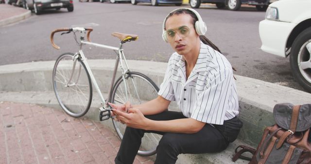 Young man sitting in a casual style with a bicycle nearby, wearing headphones and glasses, looking at camera, connected with a digital device. Perfect for lifestyle blogs, urban and digital technology advertisements, youth culture promotions, and modern fashion catalogs.