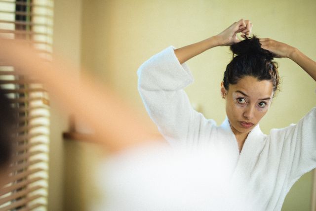 Biracial woman with vitiligo tying her hair in front of a mirror while wearing a bathrobe. Ideal for use in content related to self-care, beauty routines, spa experiences, body positivity, and wellness. Can be used in blogs, articles, and advertisements promoting skincare, personal care products, and holistic health.