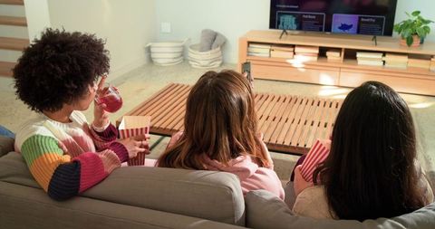 Multiracial friends sitting on sofa watching streaming tv together and sharing popcorn