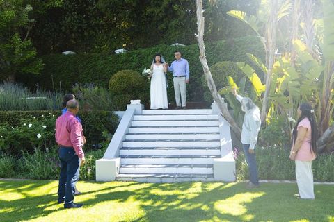 Bride and groom posing on white sunlit garden steps while guests capturing bouquet moment