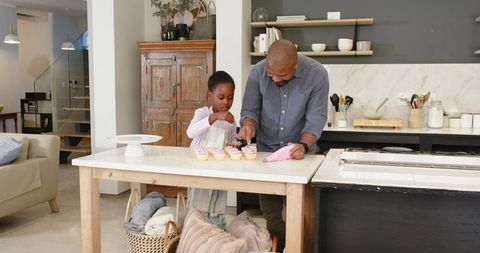 African american father and daughter decorating cupcakes together in modern kitchen