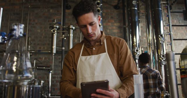 A young male worker wearing an apron is inspecting a digital tablet in an industrial brewery. The background consists of various brewing equipment, showcasing the brewing process. This visual can be used in content about modern brewing techniques, technology integration in industries, or for articles related to the brewing industry.