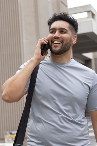Young african american man talking on phone smiling with shoulder bag urban concrete facade
