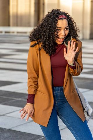 Young woman waving while talking on smartphone in sunlit urban plaza, casual style
