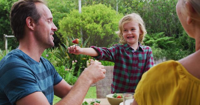 Smiling caucasian boy feeding his father a strawberry during family meal in garden. family celebrating eating outdoors together.