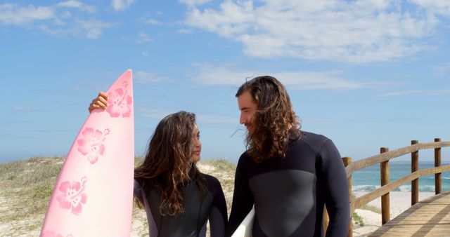 Young couple holding surfboards and walking on a beach boardwalk, dressed in wetsuits. Ideal for promoting beach activities, surfing vacations, adventure sports, and outdoor lifestyle brands.