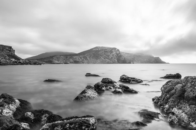 Rocky coast meeting calm sea with distant hills under a moody sky; enhanced in black and white. Ideal for decorating offices, portraying serenity and tranquility. Suitable for travel blogs, meditation backgrounds, and nature documentaries.