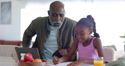 Senior man mentoring young girl with homework at sunlit kitchen table, tablet and fruit