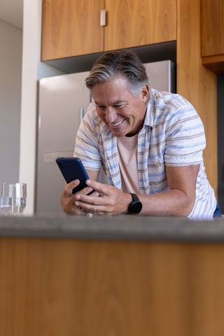 Mature man smiling while checking smartphone and wearing smartwatch in modern kitchen