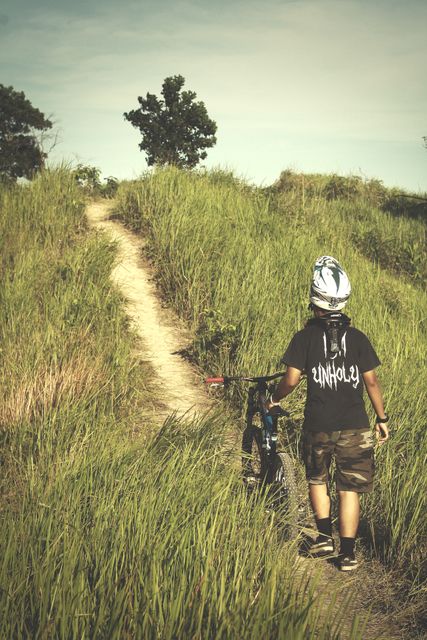 A young boy pushing his mountain bike up a narrow dirt path on a grassy hill under a clear sky. He wears a helmet for safety and sports a T-shirt and shorts, indicating summer weather. This depicts outdoor adventure, youth activity, and exploration. Represents themes of determination, fitness, and childhood fun. Ideal for use in marketing materials for outdoor gear, children's activities, adventure tours, or health and fitness campaigns.