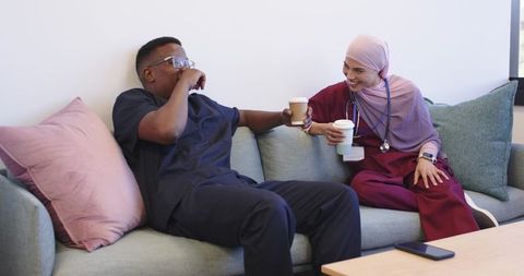 Diverse healthcare colleagues in scrubs sharing coffee and laughing on lounge couch