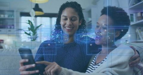 Two women sharing smartphone with augmented reality interface on cozy living room sofa