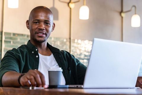 African man working remotely in cafe with laptop and coffee, smiling and focused on productivity