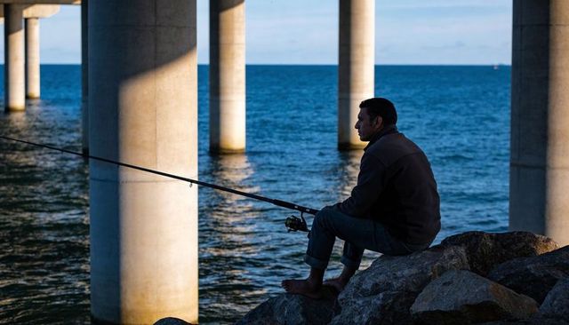 Solitary angler sitting on rocks beneath pier, casting line over coastal water at dusk