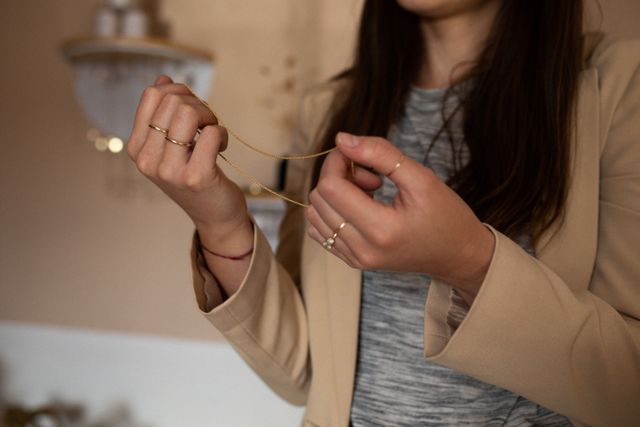 Caucasian woman holding a gold chain in a jewelry shop. She is wearing a beige blazer and several rings. This image is ideal for use in marketing materials for jewelry stores, fashion blogs, and advertisements for handmade or independent craft businesses. It conveys elegance, style, and the personal touch of handmade jewelry.