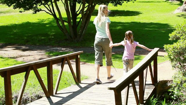 Mother and young daughter walking hand in hand on a small wooden bridge amidst lush greenery on a sunny day. Ideal for family outing themes, parenting blogs, outdoor activities, and nature articles. Perfect for promoting parks, family-friendly locations, and healthy lifestyles.