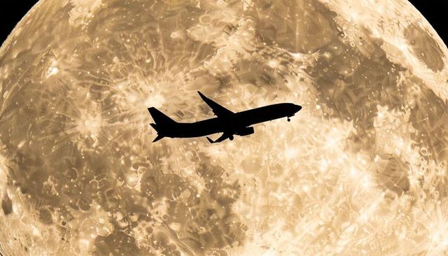 Commercial jet silhouette flying across giant lunar full moon with landing gear extended
