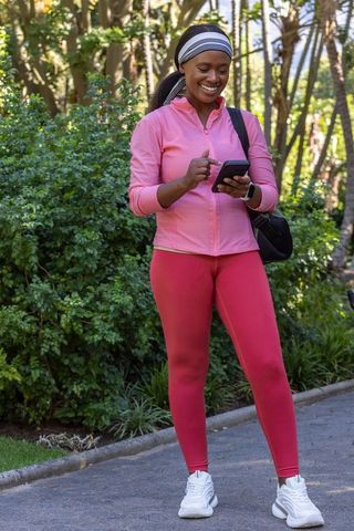 African american woman using smartphone on park walk wearing pink activewear and smartwatch