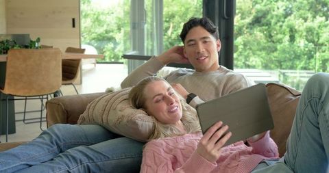 Mixed-race couple relaxing on couch sharing tablet in sunlit modern living room