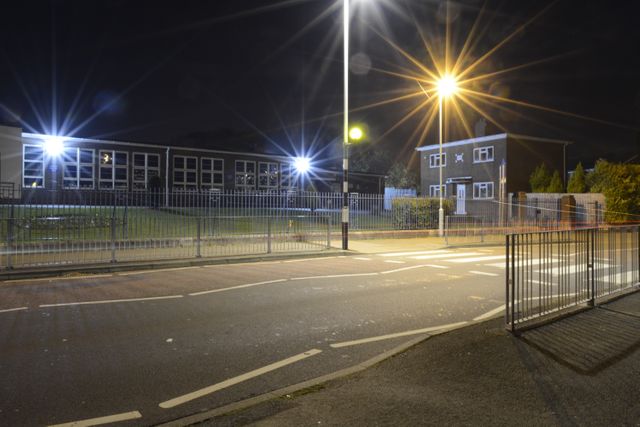 Photo of an empty street at night in a quiet residential neighborhood. Buildings are illuminated by street lights, adding a tranquil ambiance. The fencing around the area suggests a secure place. Suitable for illustrating night scenes, tranquility, residential life, and urban landscapes.