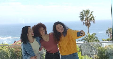 Diverse women taking selfie on coastal balcony with glass railing, ocean view, casual fun