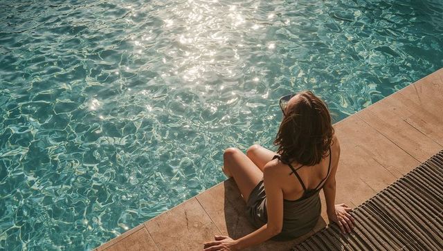 Relaxing woman sitting by sunlit turquoise pool on tiled coping wearing strappy swimsuit