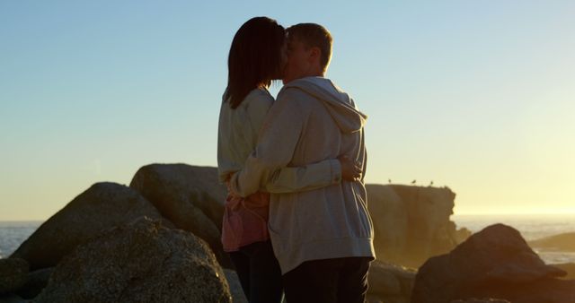 A young Caucasian couple shares a romantic moment at sunset by the sea, with copy space. Their silhouettes against the tranquil backdrop of the ocean create a serene and intimate atmosphere.