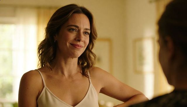 Leaning woman smiling gently in sunlit room wearing beige camisole and hoop earrings