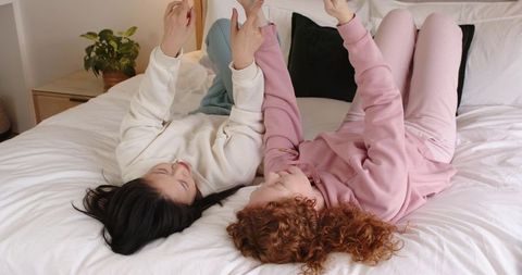 Diverse female friends lounging on bed scrolling smartphones in cozy pastel loungewear