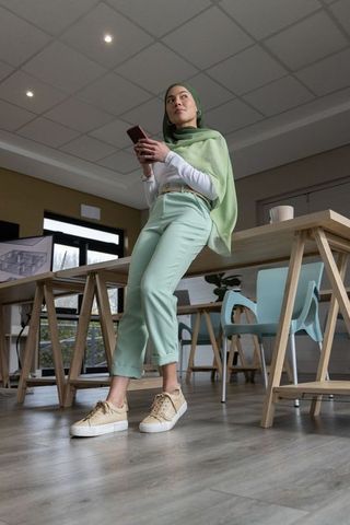 Young professional wearing green hijab leaning on desk holding smartphone at coworking hub