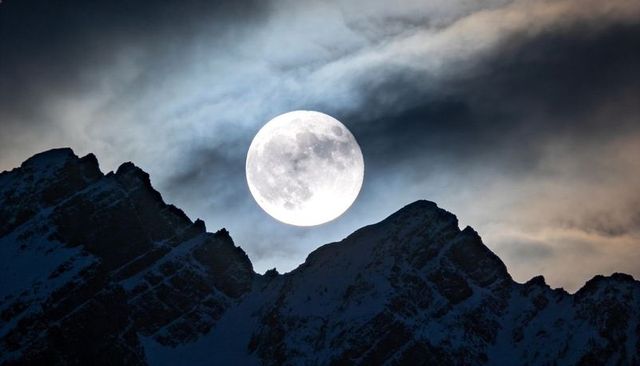 Full moon rising above jagged alpine ridge with dramatic clouds and snow-capped peaks