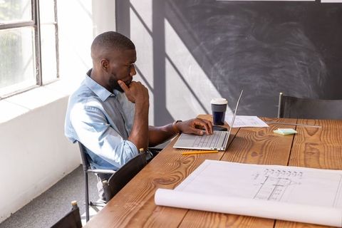 African American Architect Working on Laptop with Blueprints in Light-Filled Workspace