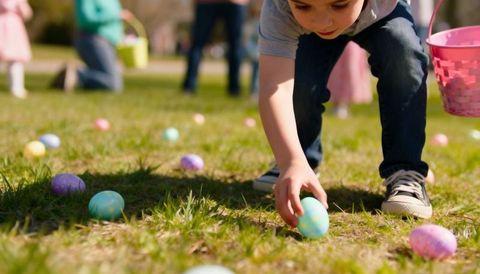 Young child collecting pastel easter eggs on sunlit grass with pink basket and group