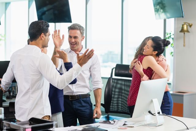 Business colleagues are seen celebrating a success in a modern office environment. They are smiling, embracing, and high-fiving, indicating a moment of achievement and teamwork. This image can be used to depict themes of corporate success, teamwork, and positive workplace culture. It is suitable for business presentations, corporate websites, and promotional materials highlighting team spirit and professional achievements.