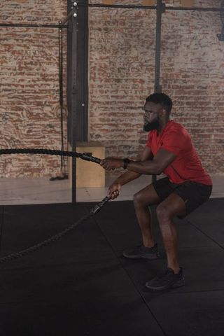 African american athlete training with battle ropes and squatting in brick industrial gym