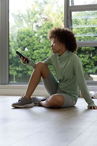 African american woman sitting on floor holding phone glancing outside in sage activewear