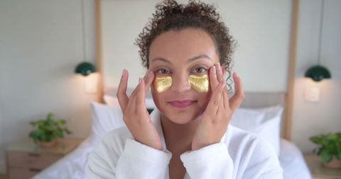 African american woman applying gold under-eye hydrogel patches in white robe for self-care