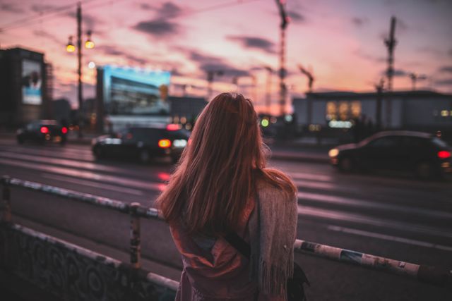 Woman with shawl standing on urban bridge, looking at cars during sunset. Ideal for depicting urban lifestyle, traveling, evening serenity, and introspective moments. It could be used in projects related to city life, travel blogs, mental health, and relaxation.