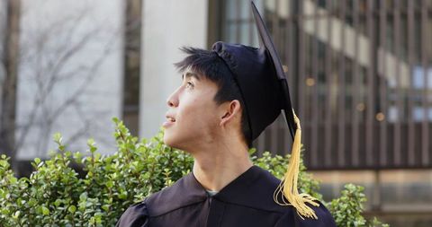 Asian male graduate looking upward in cap and gown on university campus courtyard