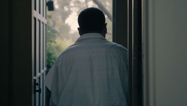 Man standing in open doorway looking out to garden from narrow hallway at dusk