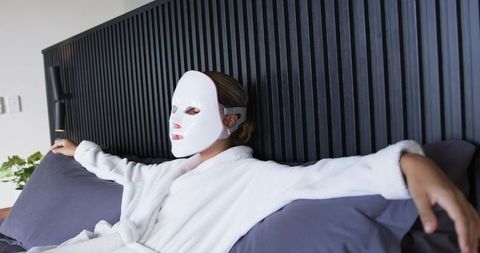 Woman reclining in bed wearing led facial mask and white robe nighttime skincare routine