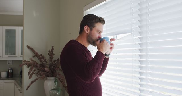 Image of thoughtful caucasian man drinking coffee and looking outside window. Lifestyle, relax, and spending free time at home concept.